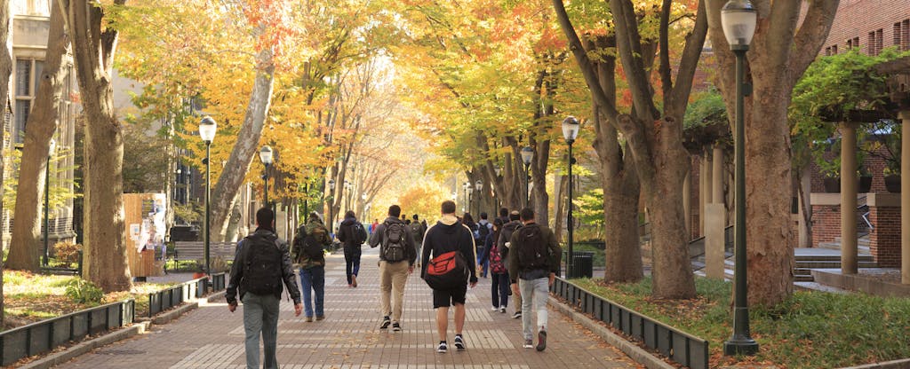 Students walk along a paved campus walkway beneath brightly colored fall foliage.