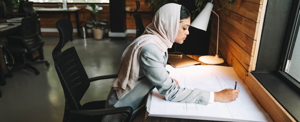 An architect wearing a hijab sits at a desk while drafting on a large piece of paper.