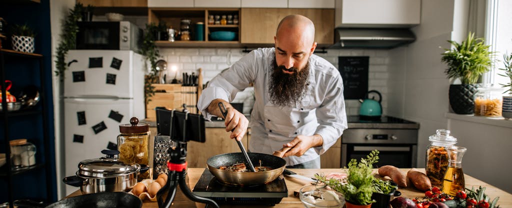 A bearded person uses their smartphone and a tripod to film a cooking video in their kitchen.