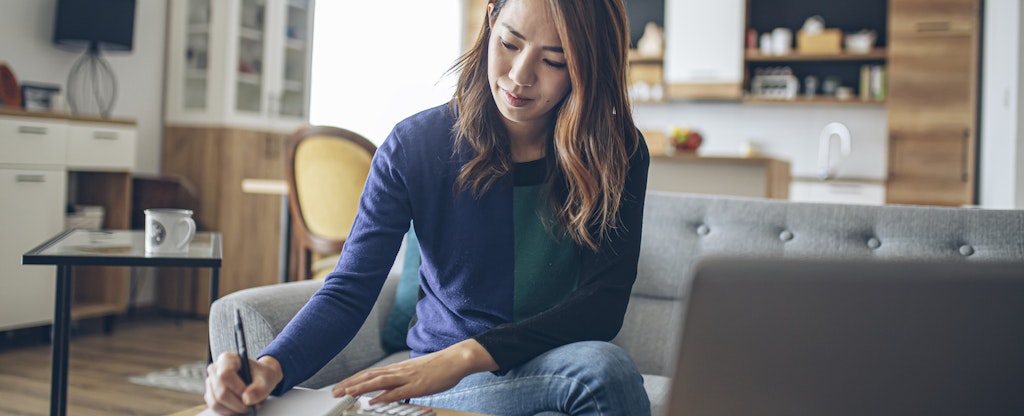 A person seated on a sofa leans over their coffee table to take notes while using a laptop to check the status of their investments.