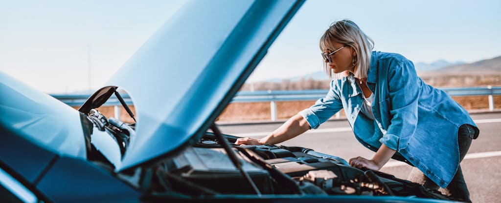 Woman looking under the hood of her car while broken down on the side of the road