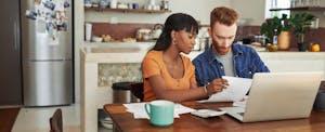 A couple sit at their kitchen table reviewing their finances using a laptop and setting long-term financial goals.