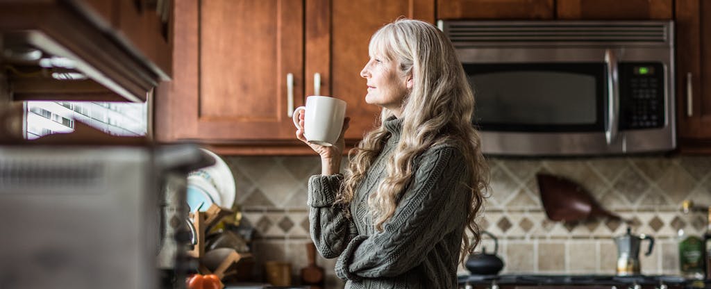 Woman standing in her kitchen, looking thoughtful as she drinks a cup of coffee