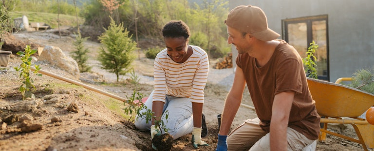 A couple kneel side by side in their backyard while planting new plants as a part of a landscaping project