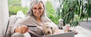 A smiling woman with grey hair and glasses holds her credit card and uses a laptop while her dog sits in her lap.