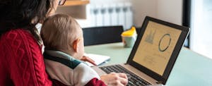 A parent seated at a desk with a baby on their lap uses a laptop to review their investments
