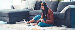 Young woman sitting on her living room floor, cup of coffee in hand, laptop open, working on managing her living expenses