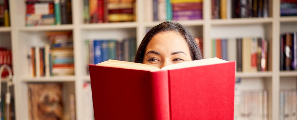 Woman peering over the top of a red book open in front of her, with a bookshelf behind her