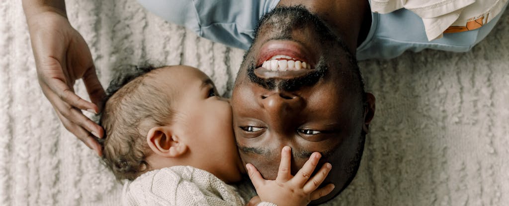 Young black father facing up on a bed, with his infant child smiling and touching his face