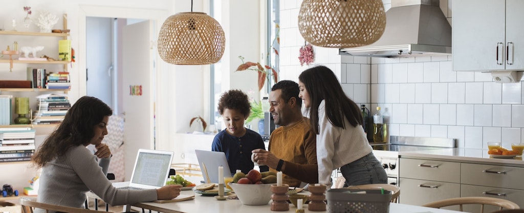 Parents sitting at the kitchen table, laptops open, working while their kids sit with them