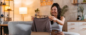 A smiling woman pulling a new shirt out of a package while sitting on a couch.