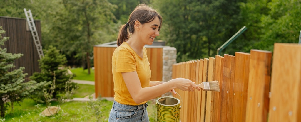 A smiling woman in a yellow shirt paints a new wooden fence that surrounds her yard.