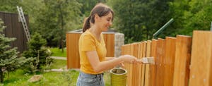 A smiling woman in a yellow shirt paints a new wooden fence that surrounds her yard.