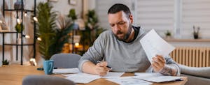A man with visible tattoos on his forearm sits at a dining room table while reviewing mortgage refinance documents.