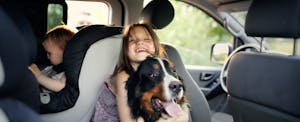 Young girl hugging dog in backseat of car with little brother in car seat.