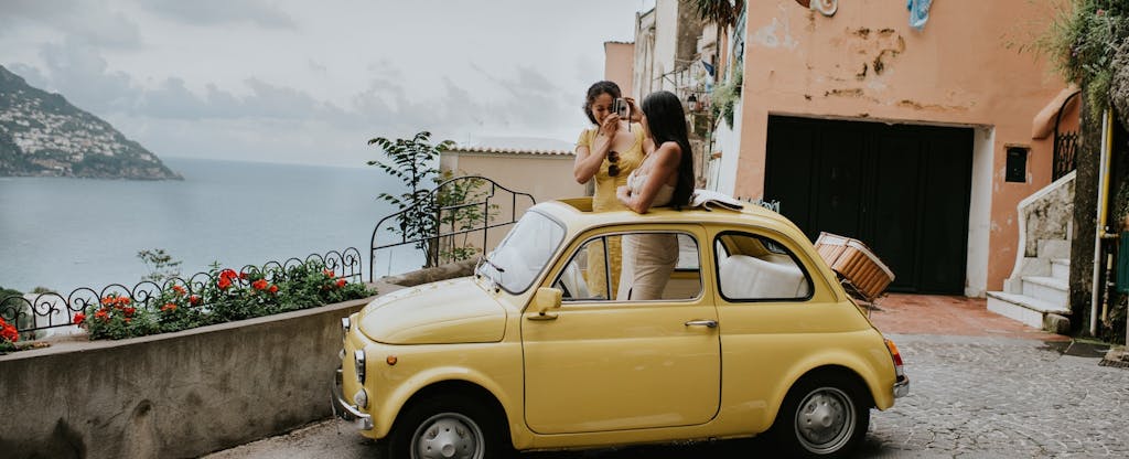 Two beautiful young woman inside a small, vintage yellow car. The top remains open. They stand up in the vehicle, through the sunroof and take photos of each other. We can see Italy's famous view of Positano in the background. Depicts a scene of experiential travel, suggesting renting a car, travel tours, European road trips and nostalgia.