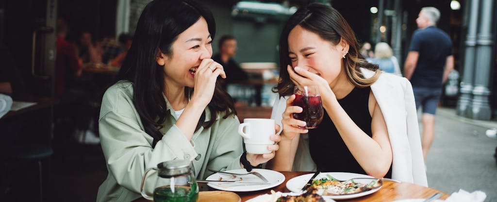 Two friends laugh with their hands over their mouths while sharing a meal at a restaurant.