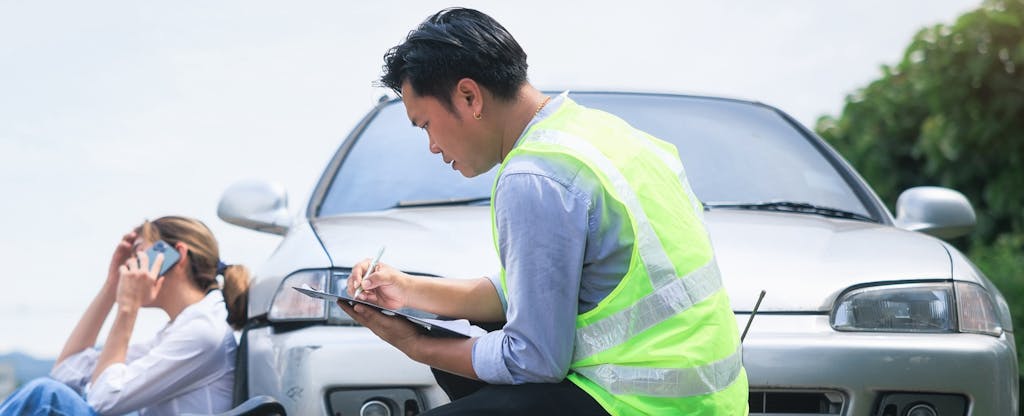 Male insurance claim adjuster examining vehicle damage while woman is sitting next to the car talking on her phone.