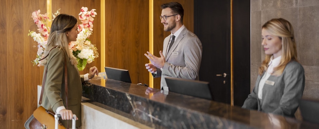 A guest stands at a hotel reception desk with their hand on the handle of their suitcase as a concierge assists them with check in