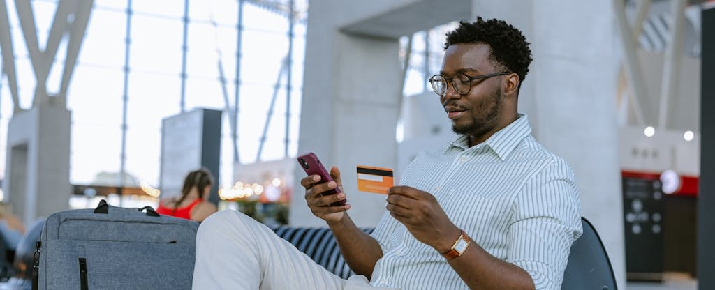 A man seated next to his luggage at an airport holds his credit card while using his cell phone.