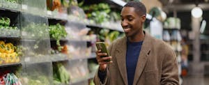 A man shopping in a grocery store, looking at his smartphone, while holding a cart loaded with items. He is near a fresh vegetable section, highlighting a modern way of shopping.