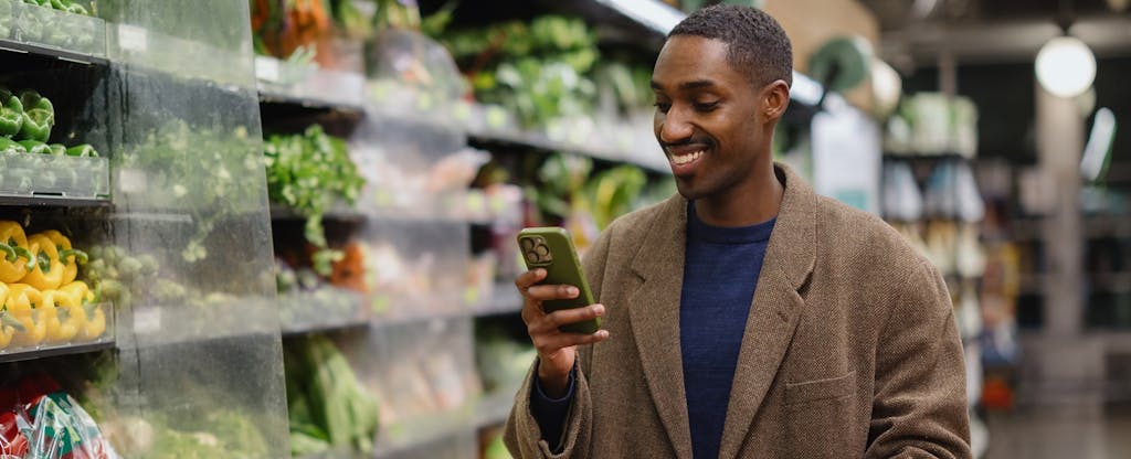 amazon-prime-visa-new-card-value A man shopping in a grocery store, looking at his smartphone, while holding a cart loaded with items. He is near a fresh vegetable section, highlighting a modern way of shopping.