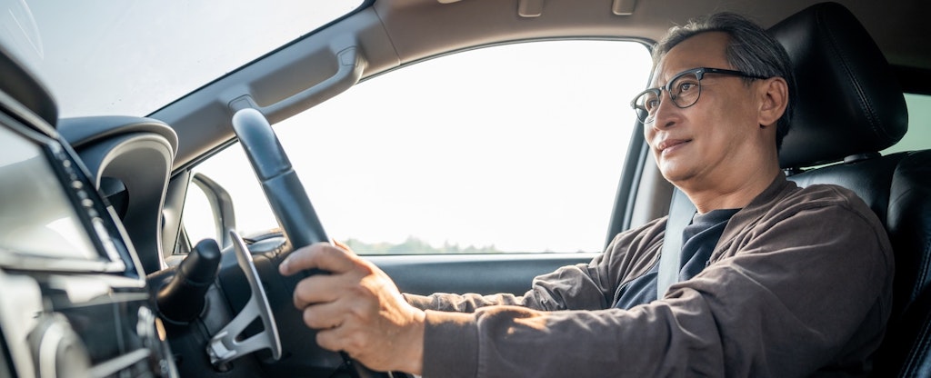 Older adult with grey hair and glasses looks ahead confidently while holding the steering wheel of his car.
