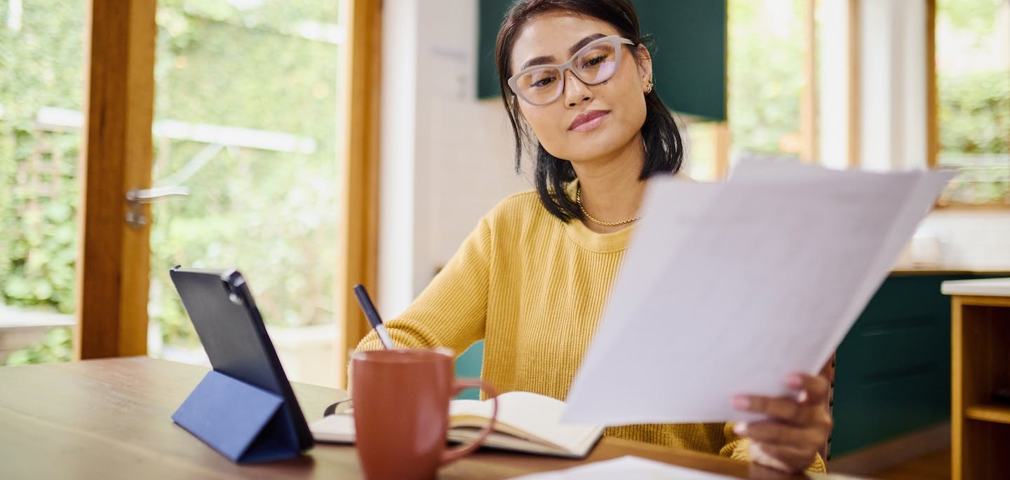 Serious Asian Woman Multi-tasking with Tablet, Notes, and Documents at Table