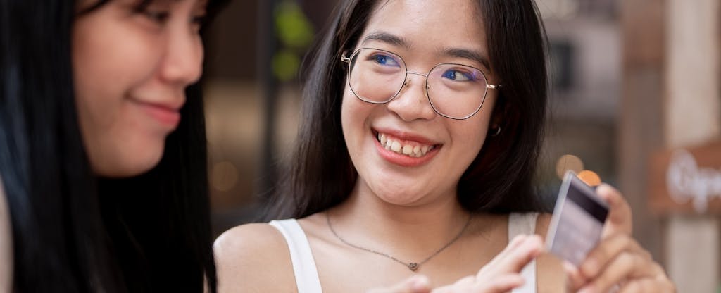Two happy Asian female friends are talking about the benefits of their credit cards at a cafe.