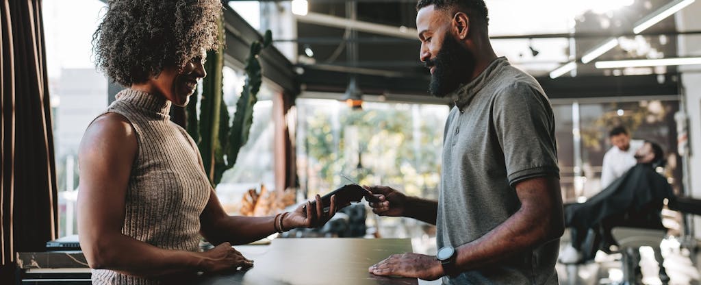 A man with a beard pays the receptionist at his barbershop using his credit card.