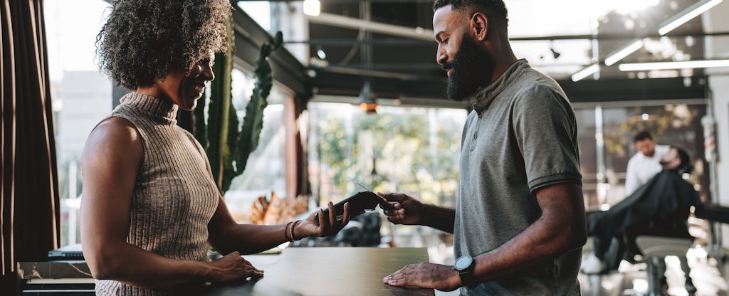 A man with a beard pays the receptionist at his barbershop using his credit card.